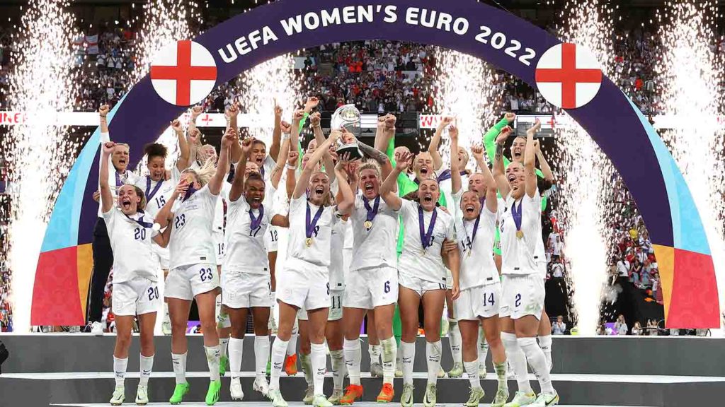 England women's national football team lifting the Euro 2022 trophy on the pitch with confetti coming down and a banner arcing over their heads.