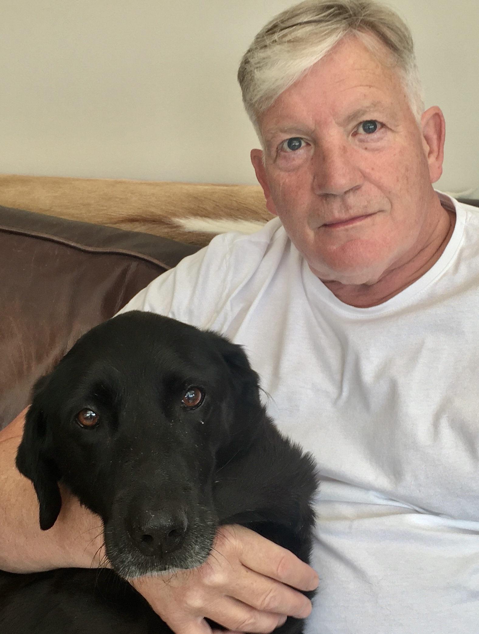 Black Labrador dog sat on sofa with her owner, a man with fair hair in a white t-shirt. Both are looking at the camera.
