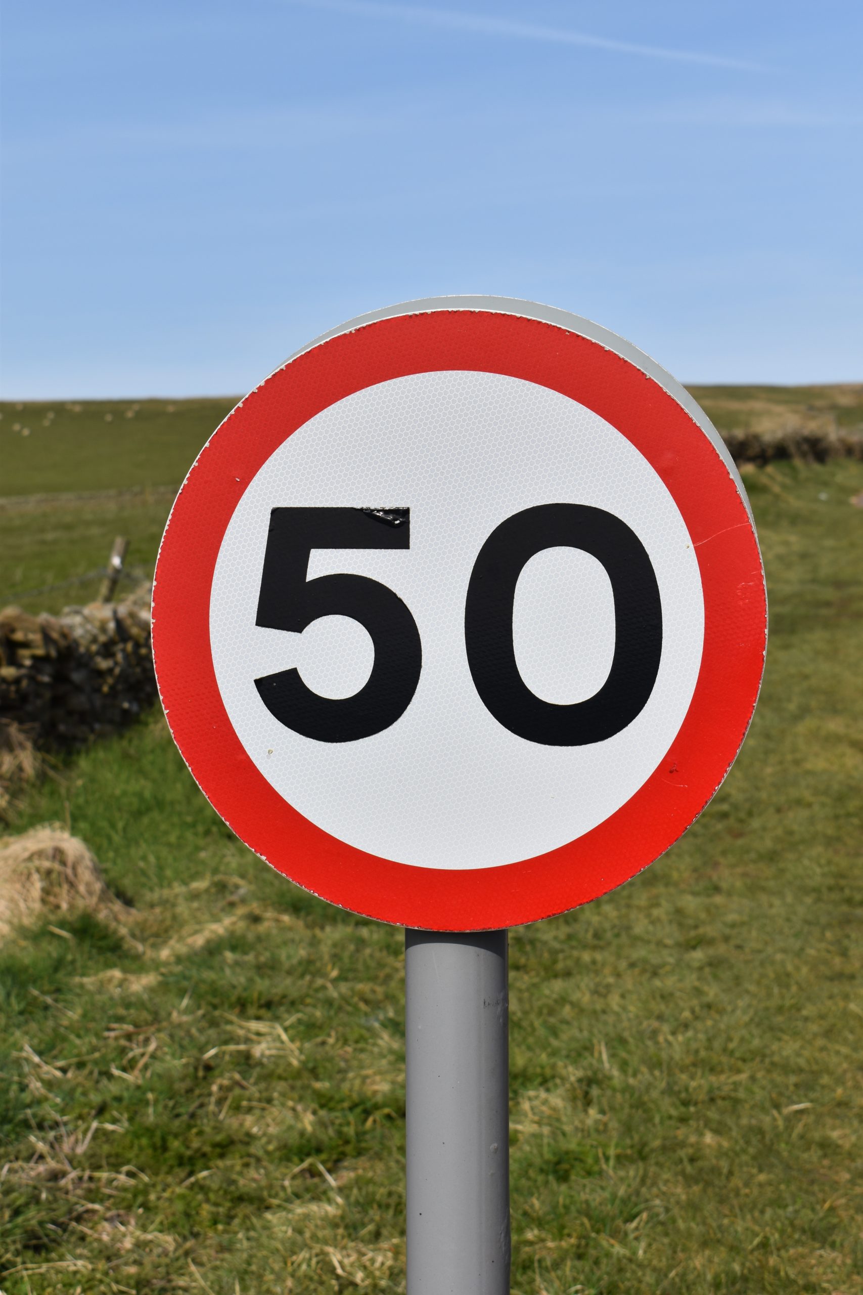 Circular traffic sign depicting 50 speed limit. Circular sign with a white background, black numbers and a red border around the edge. Countryside in the background behind the sign.