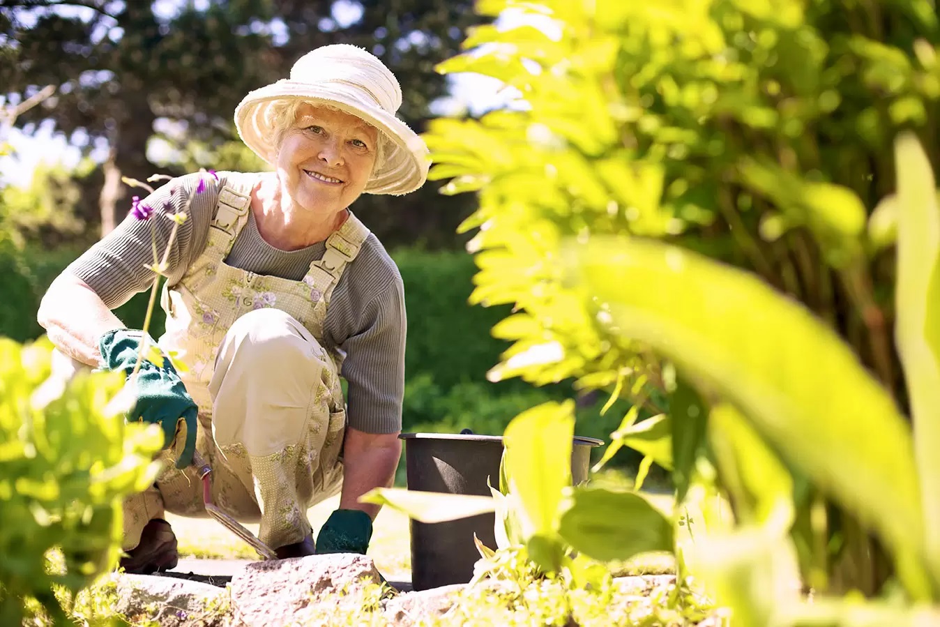 A woman is working happily in her garden. It's sunny, she's wearing a hat and she's smiling.